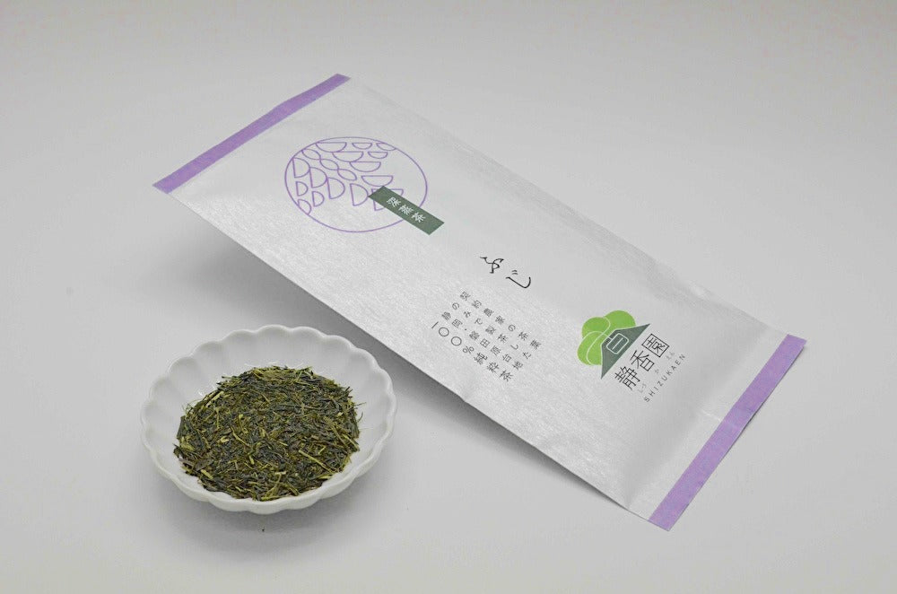 Green tea leaves in a small white dish next to a white packaging box on a light gray background