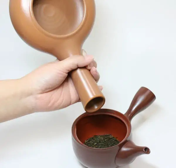 Hand pouring loose leaf green tea into ceramic teapot with wooden scoop