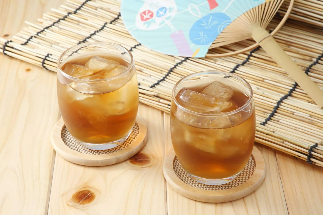 Two glasses of iced Japanese tea with ice cubes on wooden coasters, set on a bamboo mat with a decorative fan nearby.
