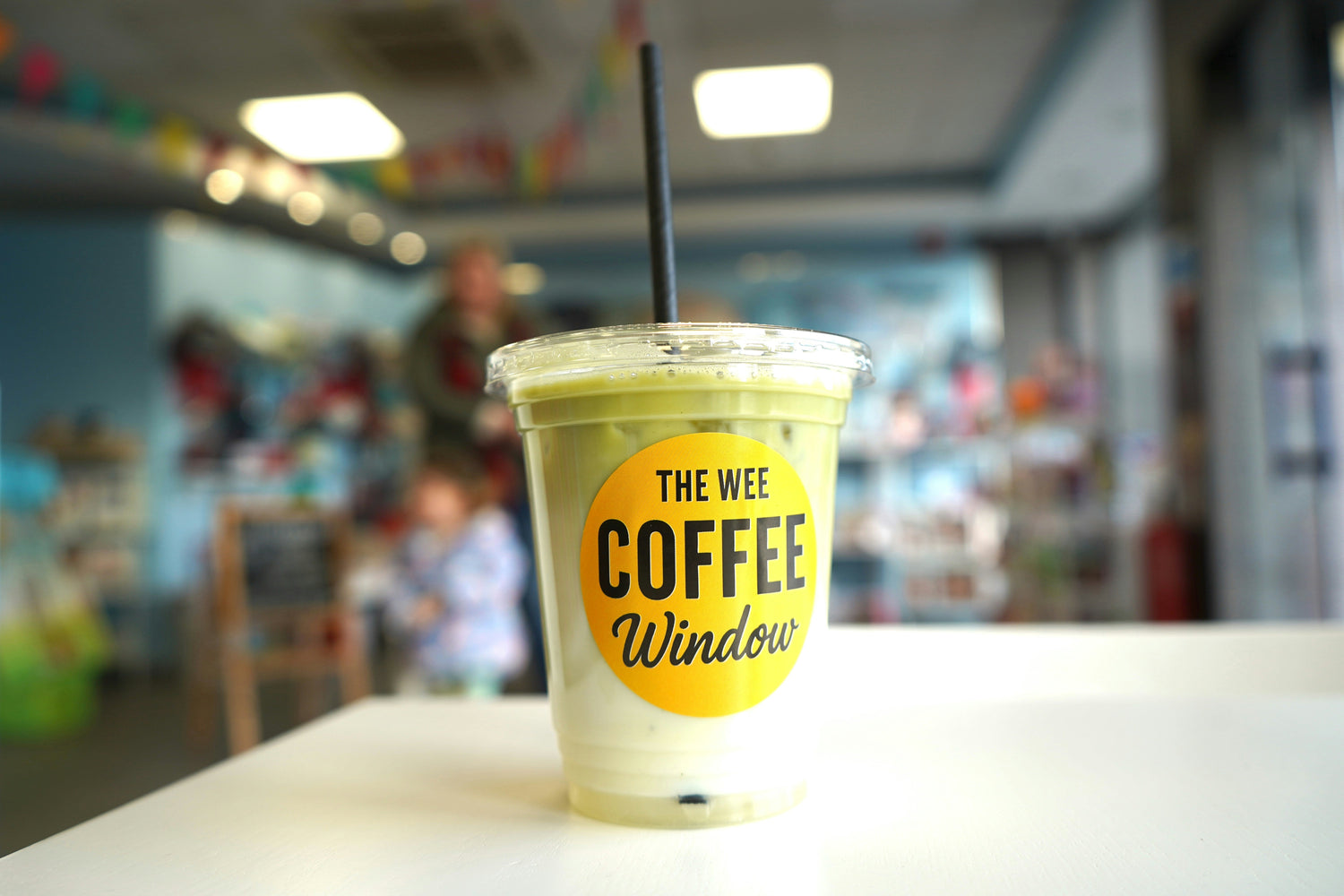 Iced matcha latte in clear cup with 'The Wee Coffee Window' label, black straw, on white table in cafe