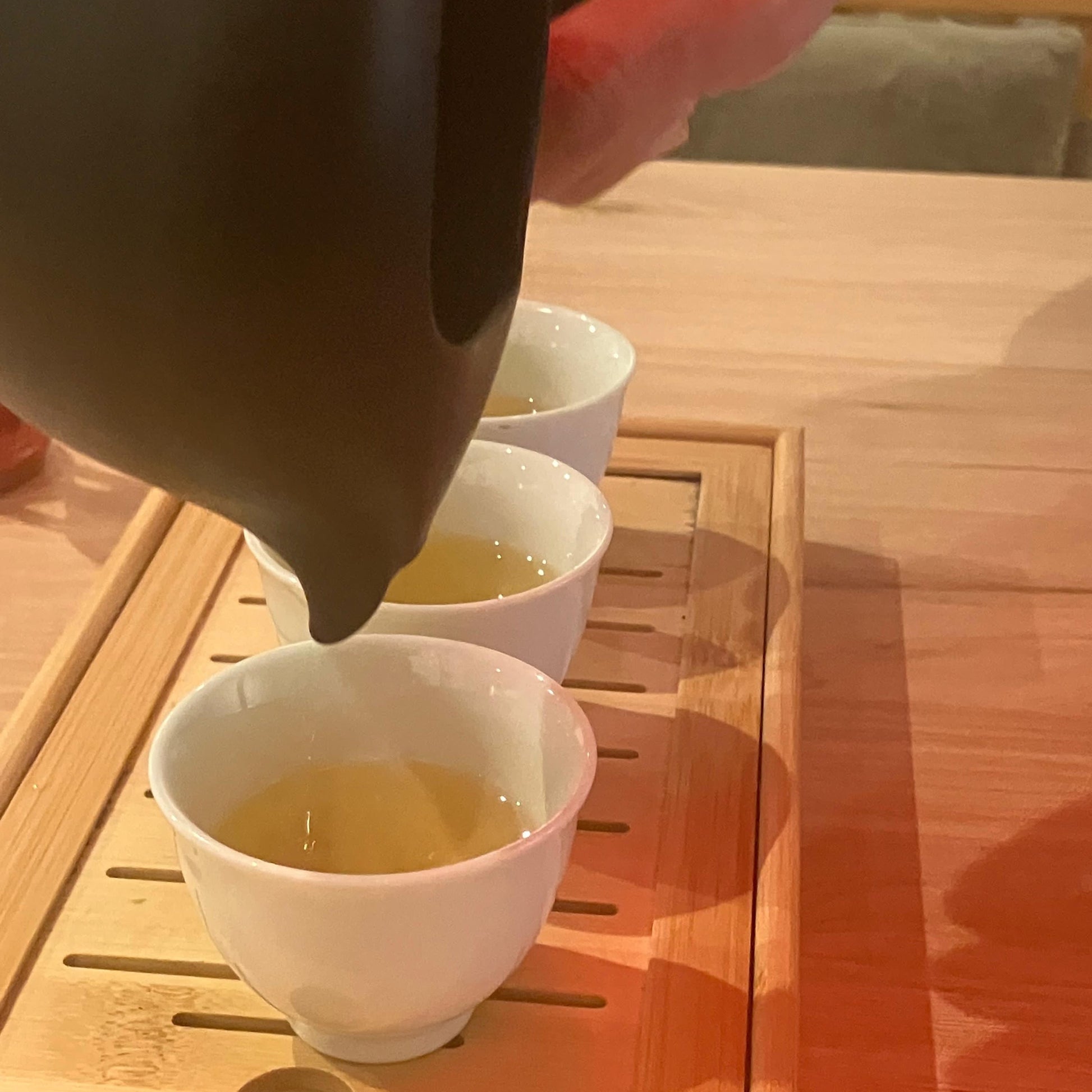 Tea being poured from a black teapot into white cups on a wooden table.