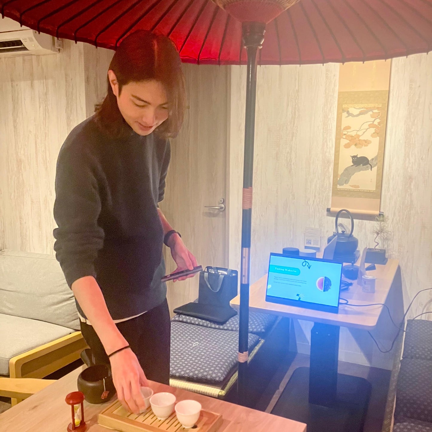 Person preparing tea in a room with a red umbrella and a computer screen in the background