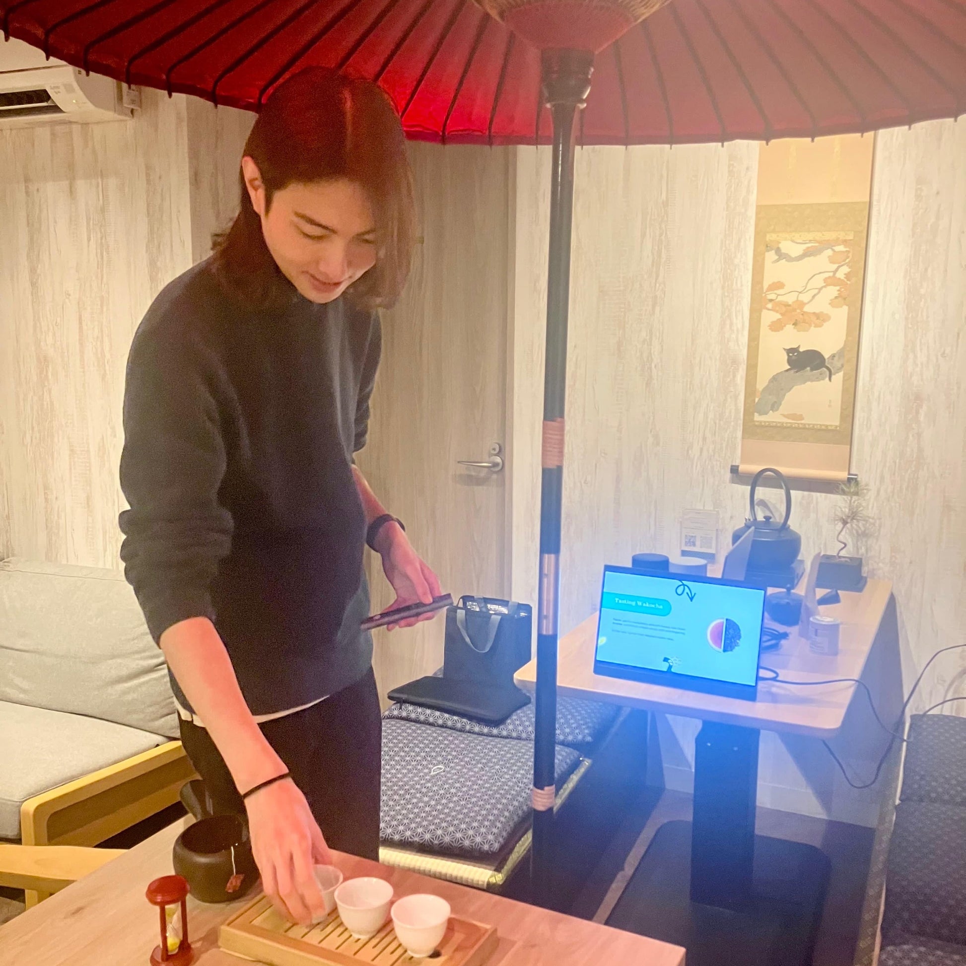 Person preparing tea in a room with a red umbrella and a computer screen in the background