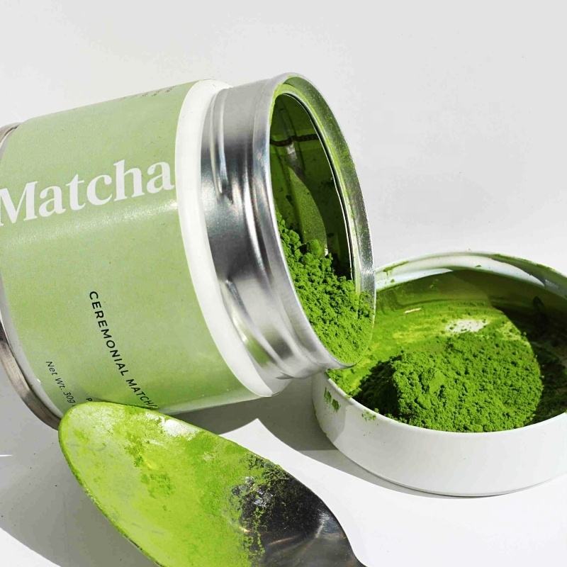 Ceremonial matcha green tea powder in open tin with spoon on white background